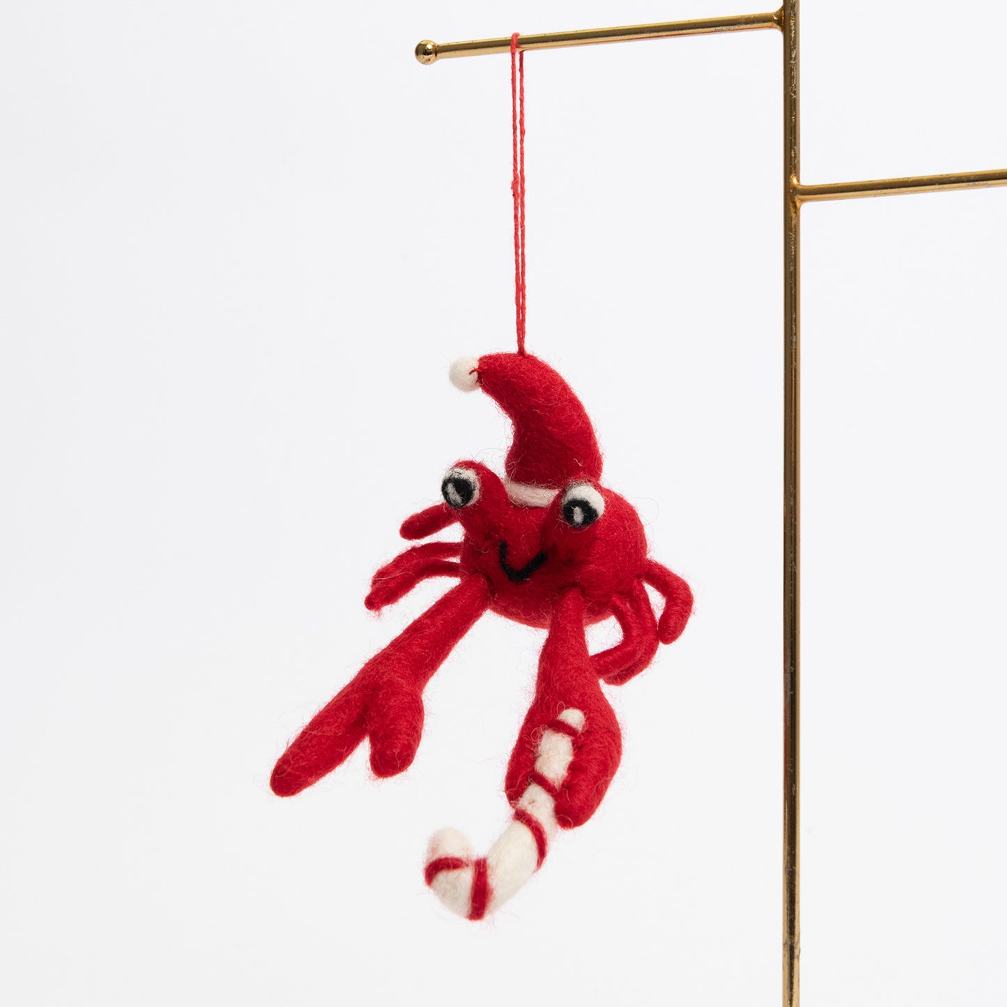A Christmas crab decoration pictured hanging from a metal rod on a white background. The felt crab is red and is wearing a Christmas hat.