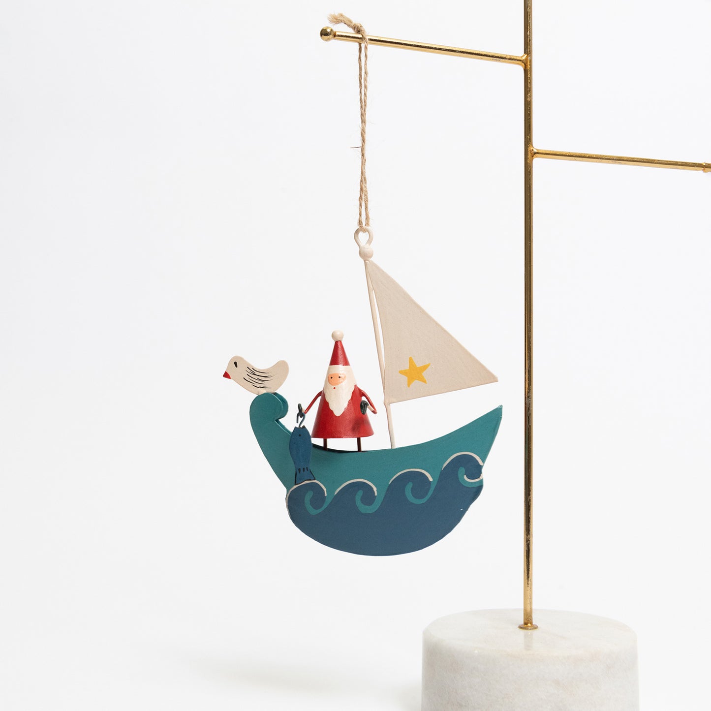 A Christmas decoration pictured hanging from a metal rod on a white background. The decoration is Santa standing on a blue sailboat with blue waves lapping up around it. There is a seagull sat on the front of the ship, and Santa is holding a fish.