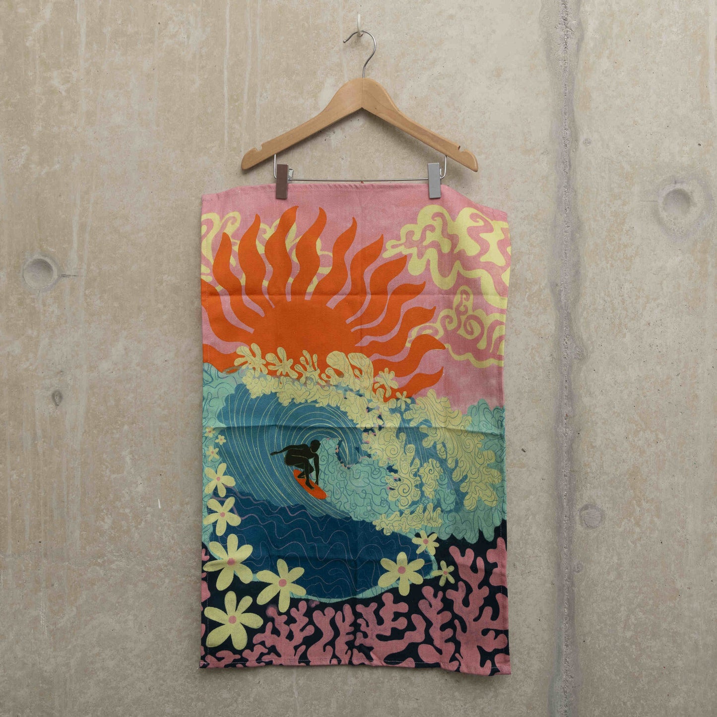 Teatowel on a hanger showing a surfer riding a big turquoise wave with flowers as foam and coral under the sea. a pink sky with a big red sun in the background.