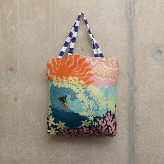 Large tote bag showing a surfer riding a big turquoise wave with flowers as foam and coral under the sea. a pink sky with a big red sun in the background.