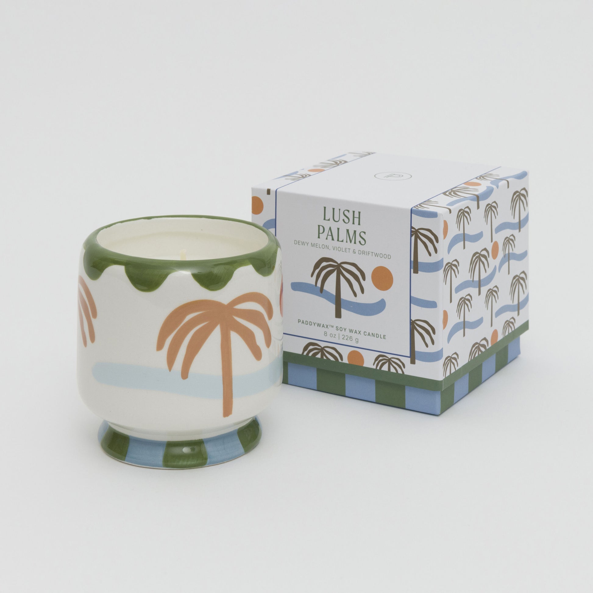 Image of soy candle in a ceramic candle holder painted with palm tree, sea and sun. Candle is placed next to the square box with matching artwork.