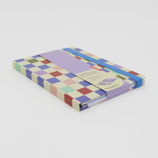 Three quarter shot of checkerboard notebook. Cream background with blue, purple, pink, green and red squares and a blue band to keep the notebook shut.