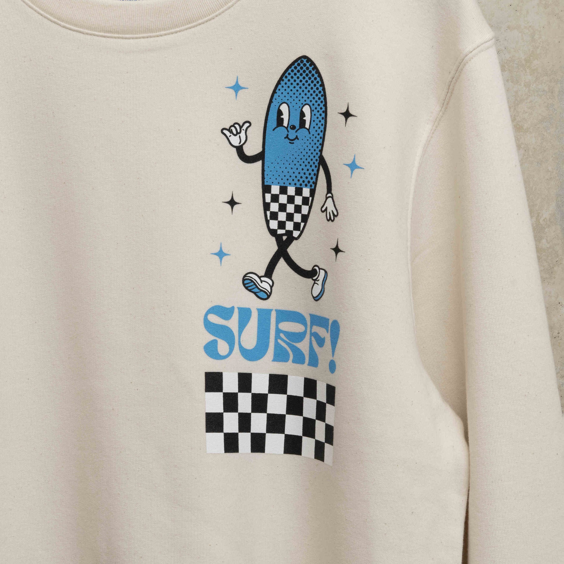 Close up of the print on left chest. Character is shaped like a surfboard in blue with black and white checkerboard design, with the word Surf underneath.
