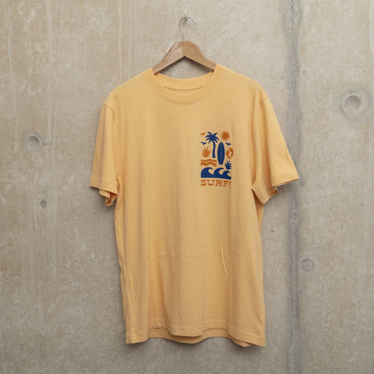 Front shot of light yellow t-shirt with surf motif on left chest. Motif shows waves, foot prints, surfboard, sun and palm tree.