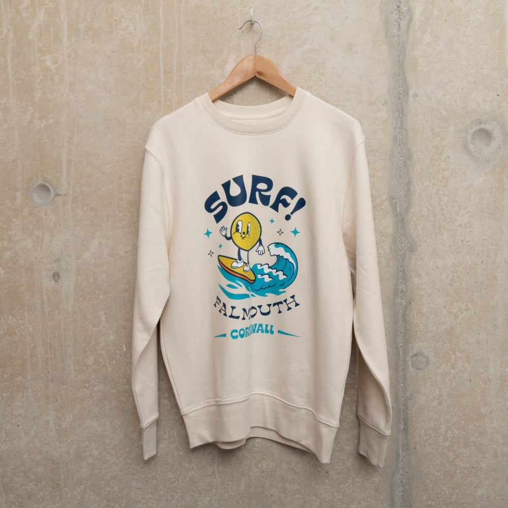 Front view of a cream sweatshirt with large motif of a character surfing a wave surrounded by the words Surf, Falmouth, Cornwall.