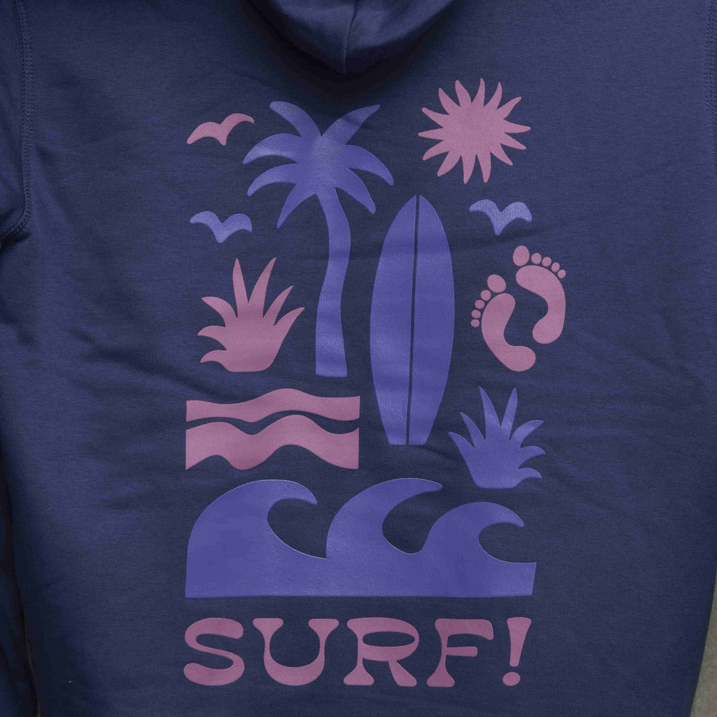 Close up of large surf motif on the back of the hoodie. Motif shows waves, foot prints, surfboard, sun and palm tree.