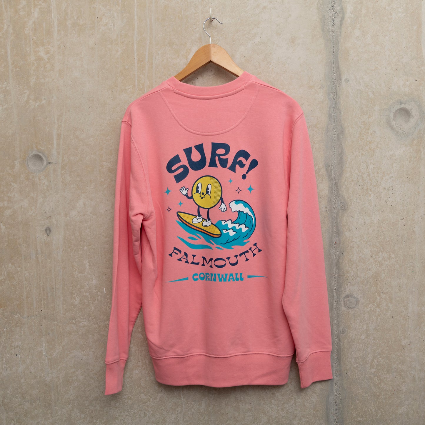 Back shot of bright pink sweatshirt with Large surf motif across the back. Motif shows round character surfing a wave with the wording SURF! Falmouth, Cornwall.