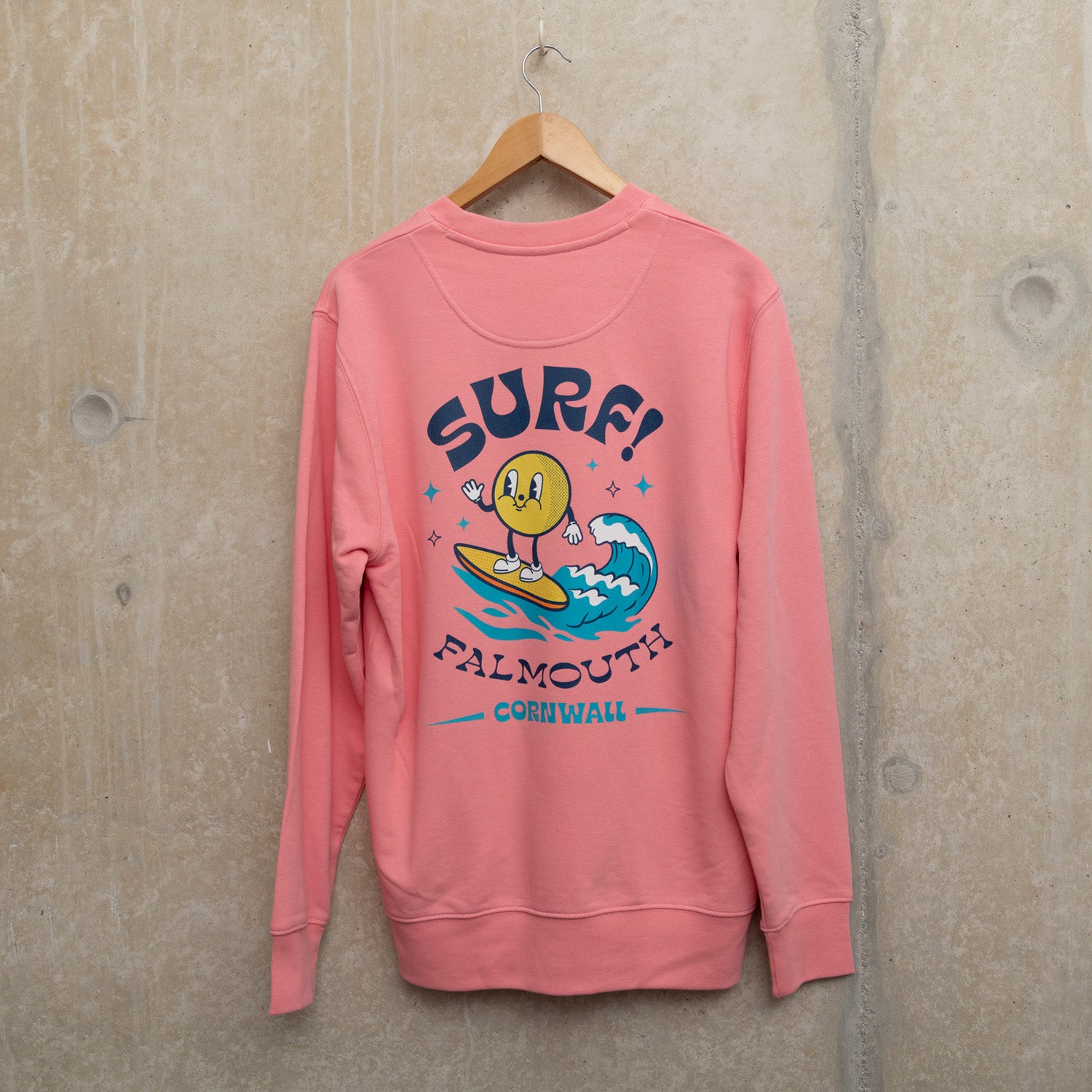 Back shot of bright pink sweatshirt with Large surf motif across the back. Motif shows round character surfing a wave with the wording SURF! Falmouth, Cornwall.
