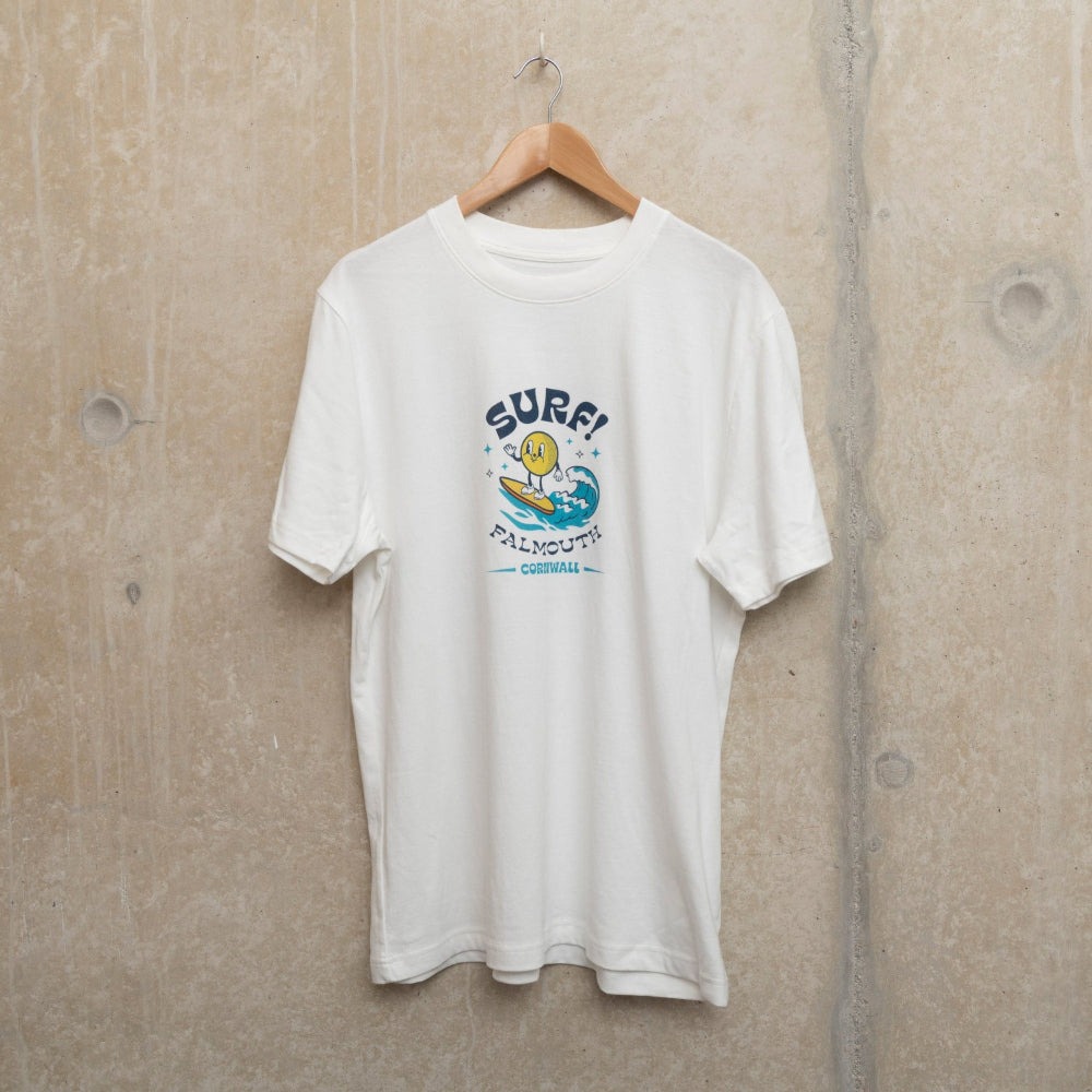Front shot of white t-shirt with medium size surf motif centre chest. Motif shows round character surfing a wave with the wording SURF! Falmouth, Cornwall.