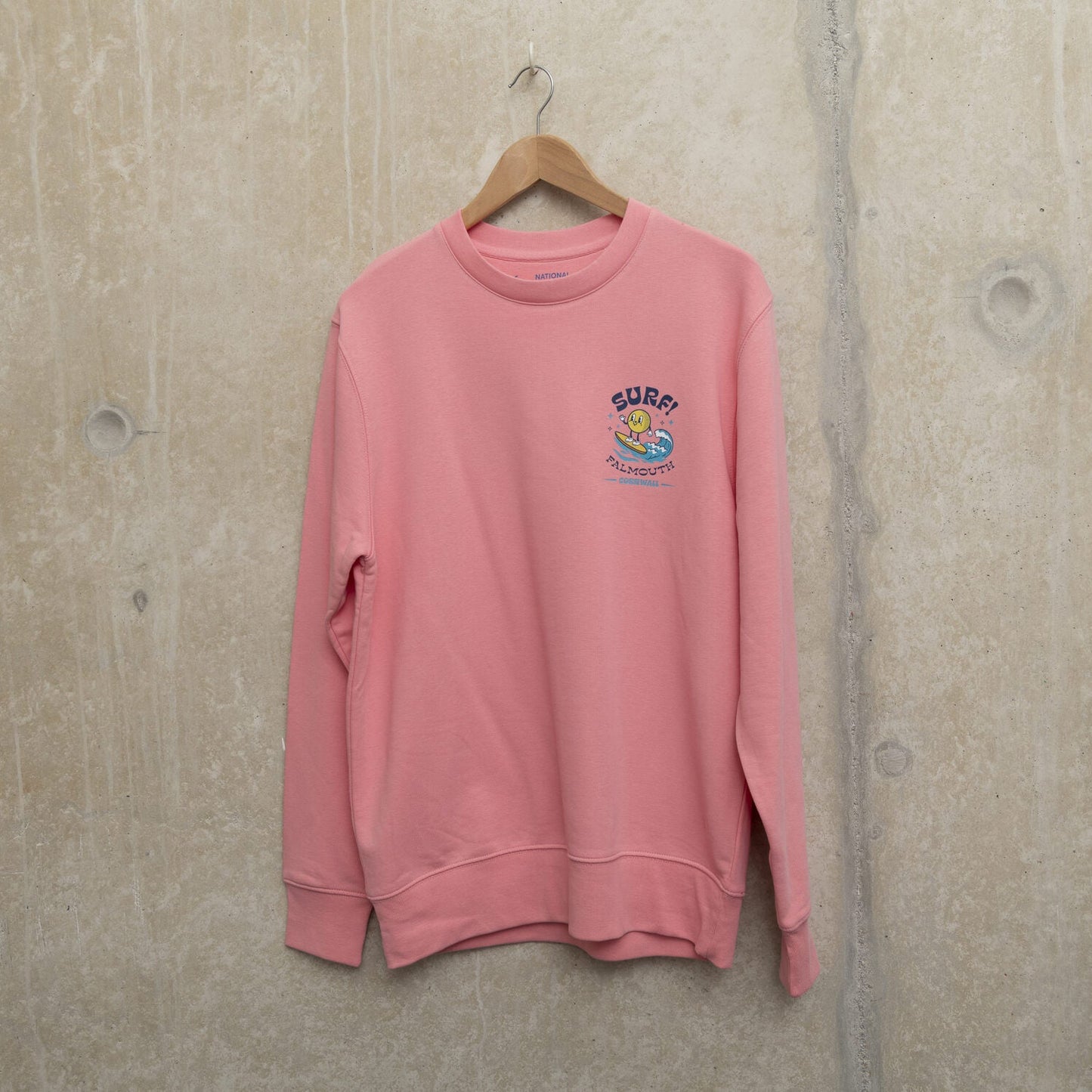 Bright pink sweatshirt with small surf motif on left chest. Motif shows round character surfing a wave with the wording SURF! Falmouth, Cornwall.