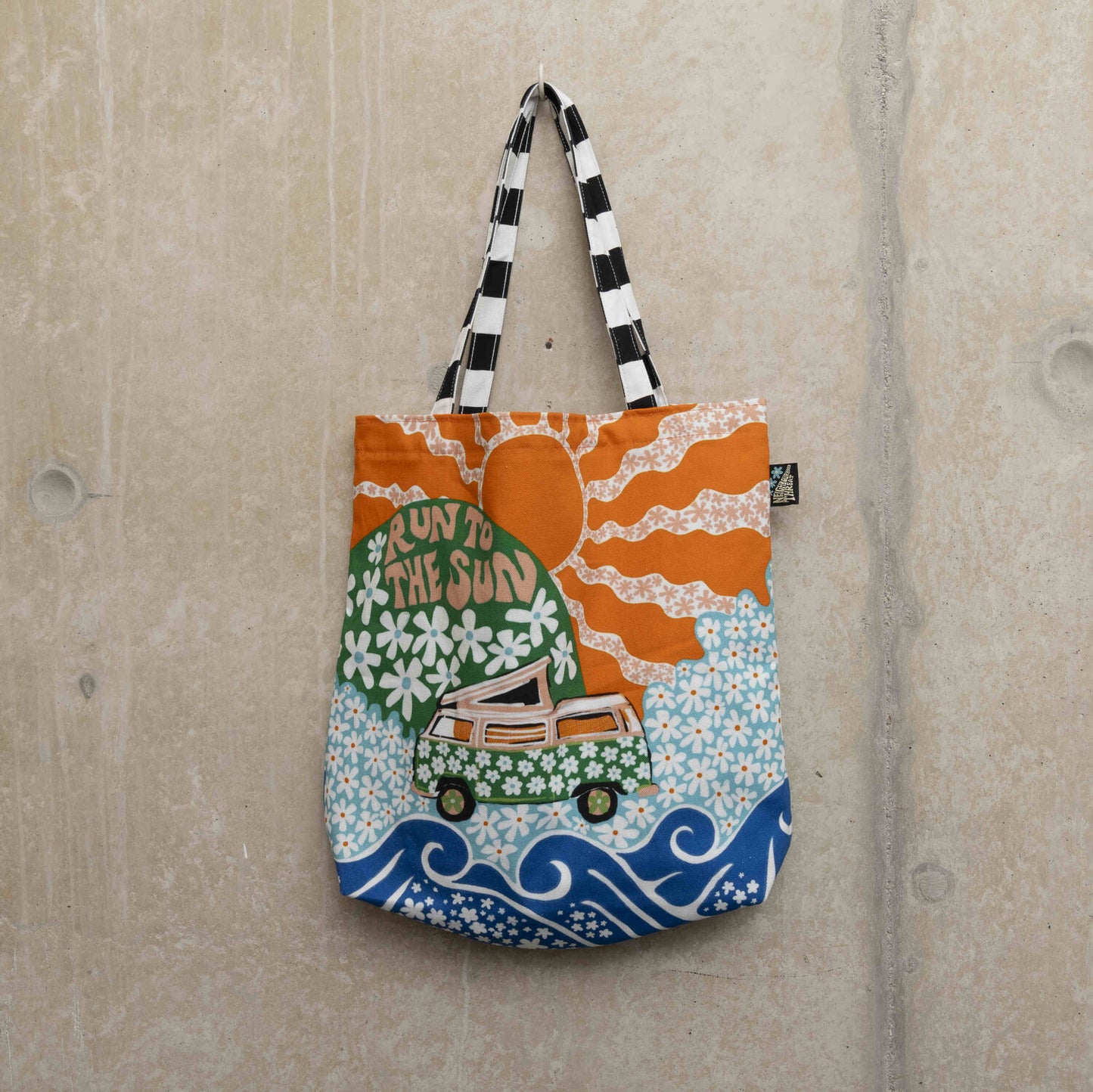 Large tote bag showing a green camper van covered in white daisies with a green hill in the background with daisies and the wording Run to the Sun in pink font. Big orange sun in the background.