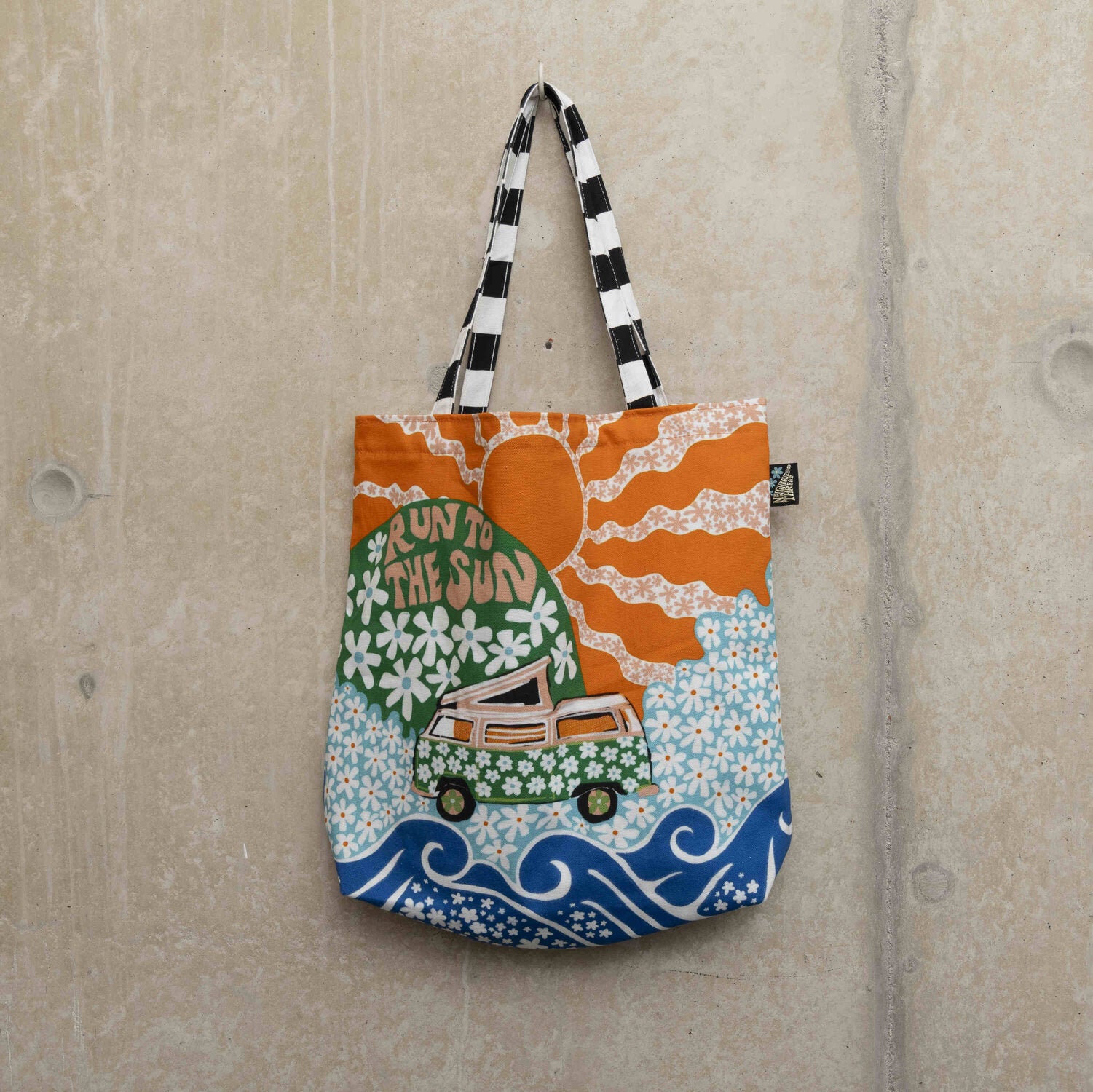 Large tote bag showing a green camper van covered in white daisies with a green hill in the background with daisies and the wording Run to the Sun in pink font. Big orange sun in the background.