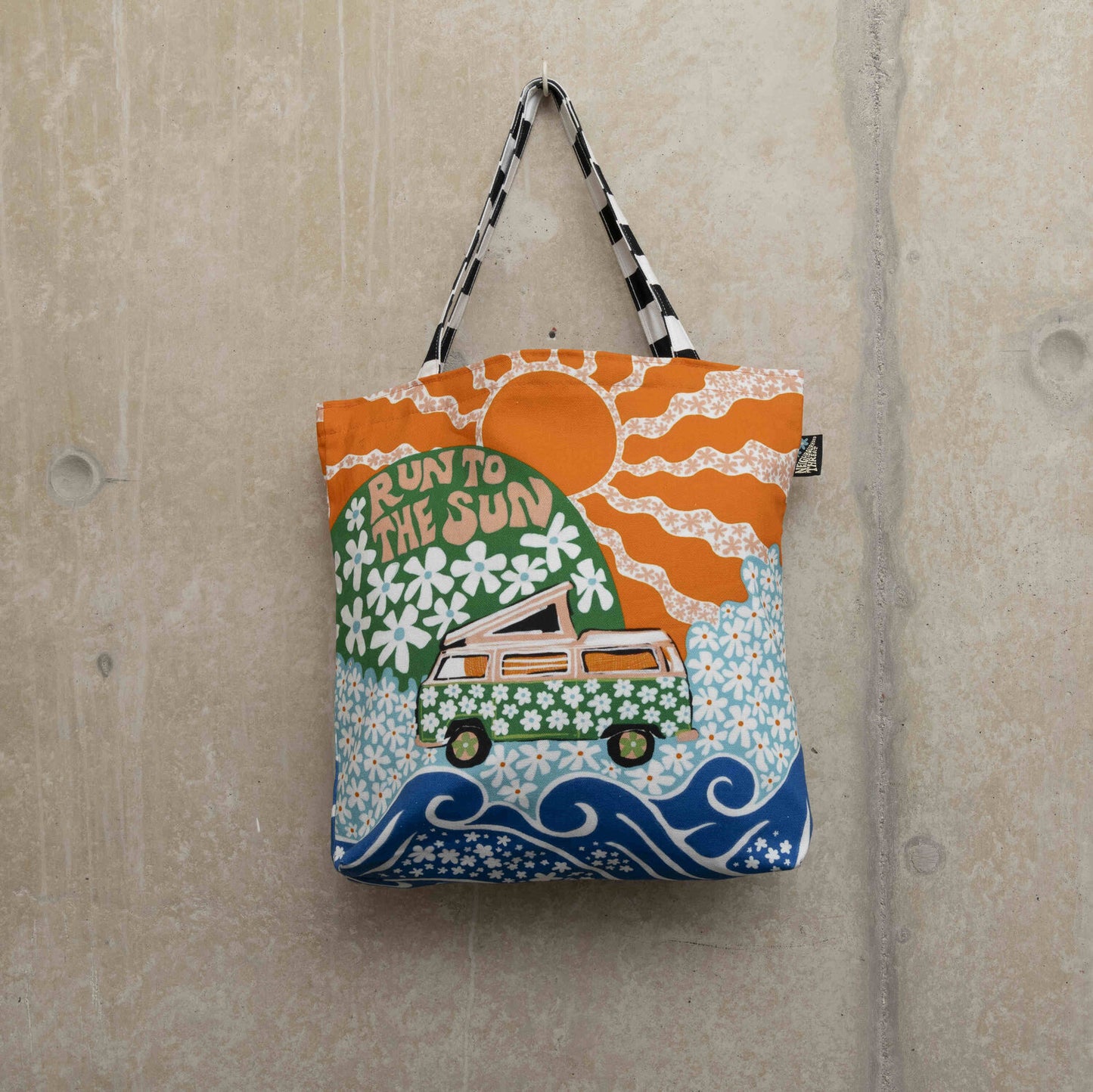 Large tote bag showing a green camper van covered in white daisies with a green hill in the background with daisies and the wording Run to the Sun in pink font. Big orange sun in the background.