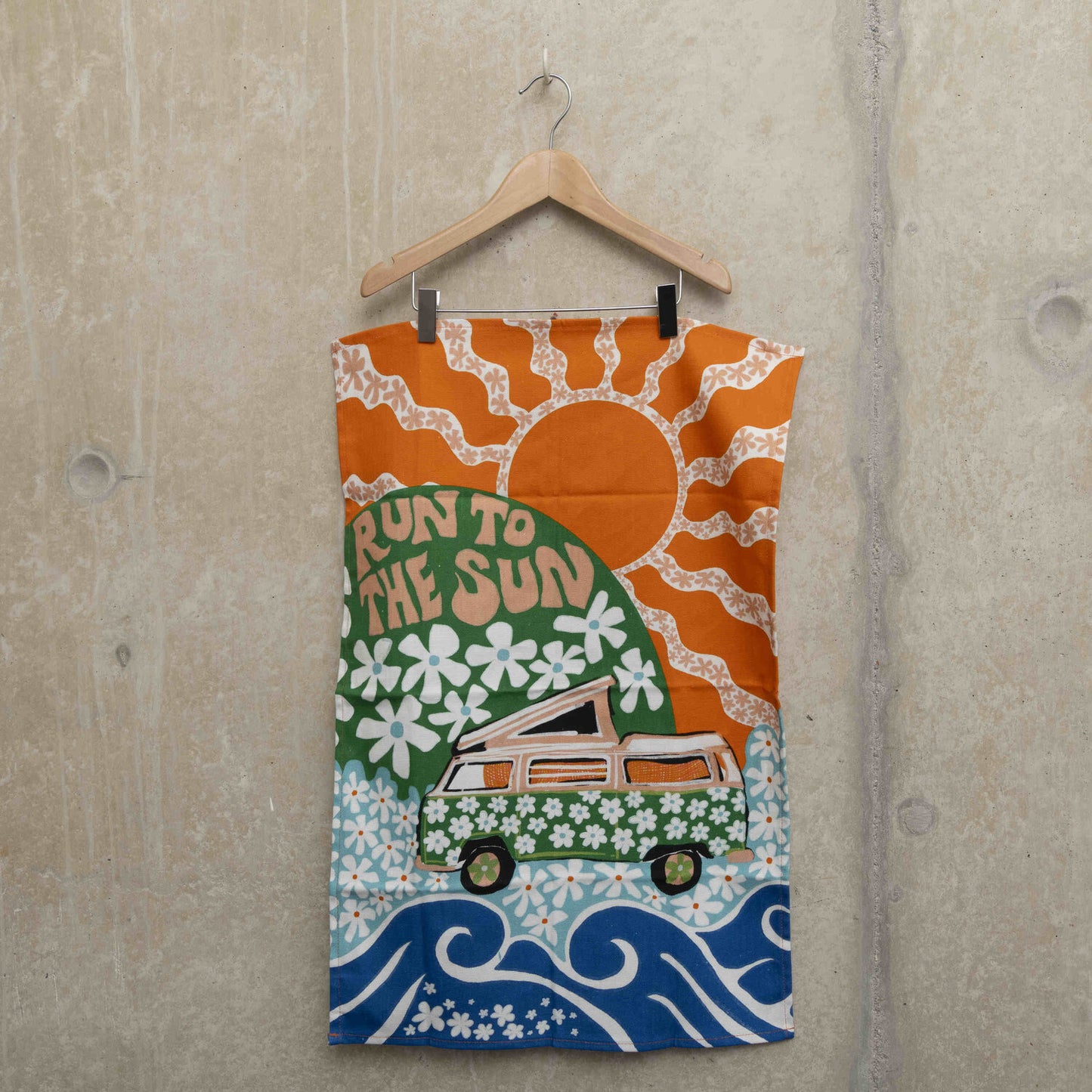Tea towel hanging on a hanger showing a green camper van covered in white daisies with a green hill in the background with daisies and the wording Run to the Sun in pink font. Big orange sun in the background.