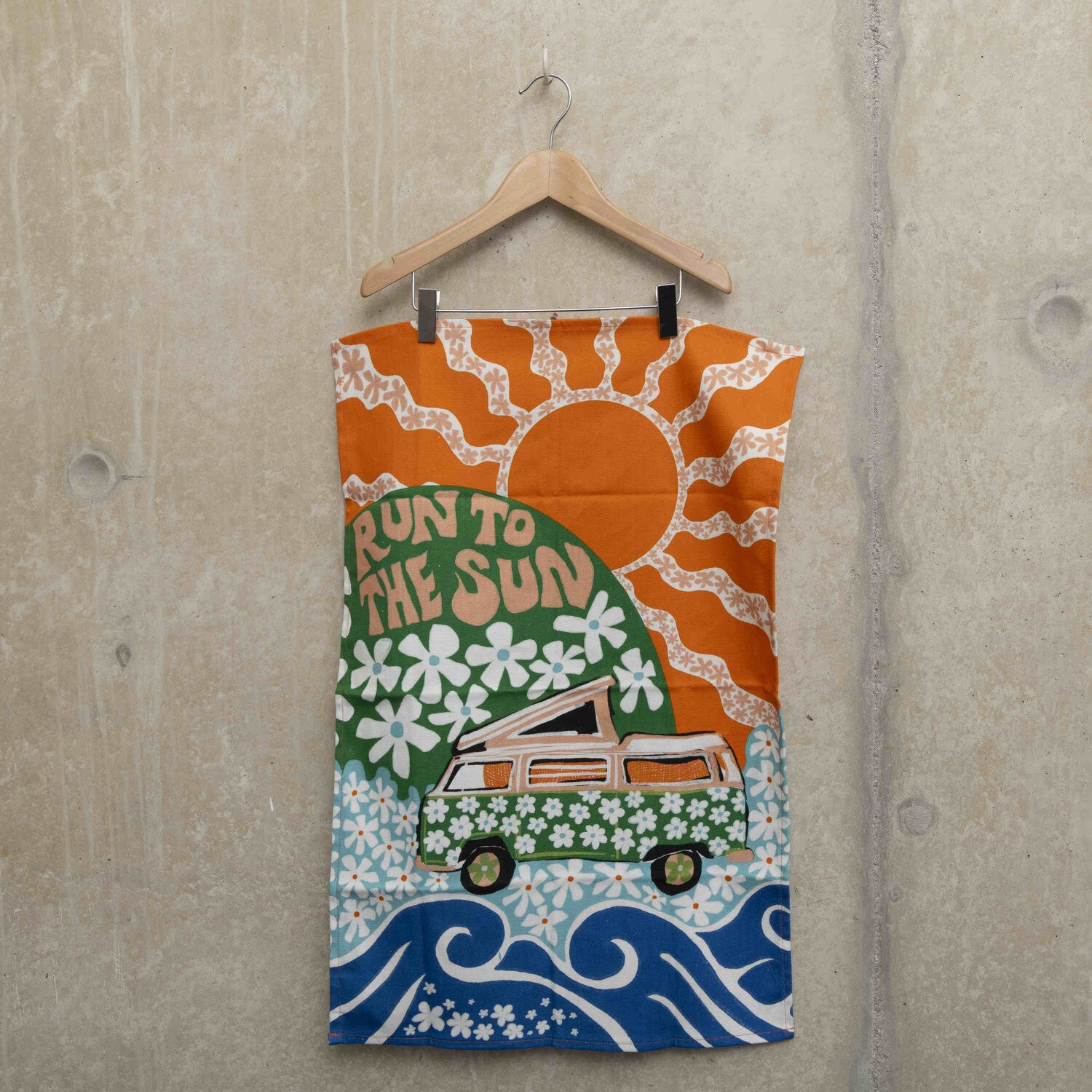 Tea towel hanging on a hanger showing a green camper van covered in white daisies with a green hill in the background with daisies and the wording Run to the Sun in pink font. Big orange sun in the background.