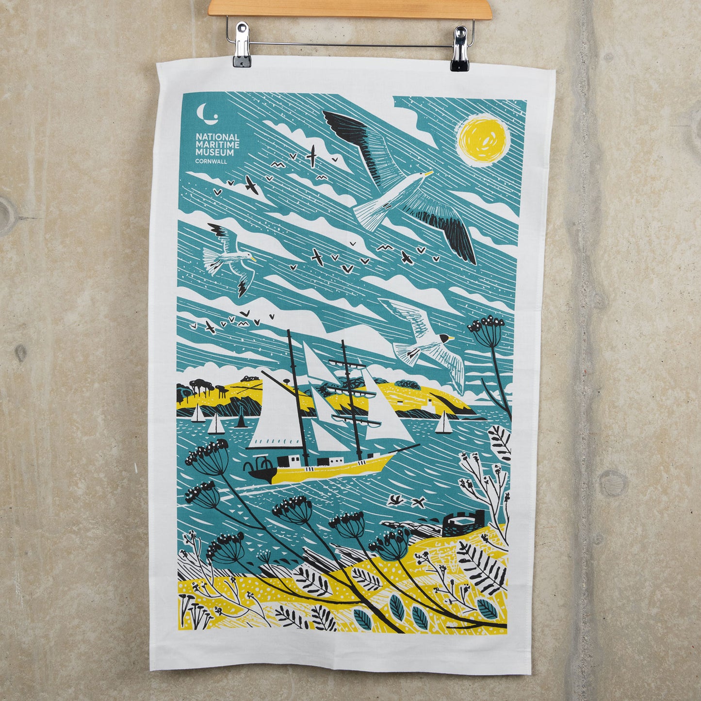 Tall ships teal towel with an illustration of a tall ship in blue, white and yellow. The tea towel is hanging from a clip hanger against a light grey background.