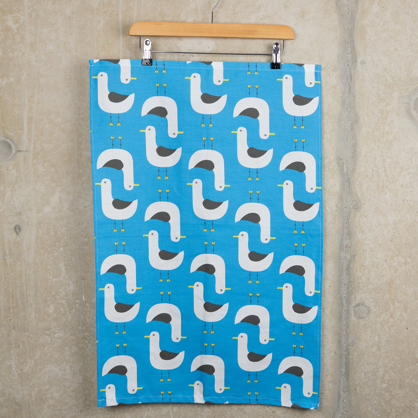 Grey and white seagull illustrations repeat across the blue seagulls tea towel. The tea towel is hanging from a clip hanger on a grey background.