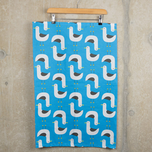 Grey and white seagull illustrations repeat across the blue seagulls tea towel. The tea towel is hanging from a clip hanger on a grey background.