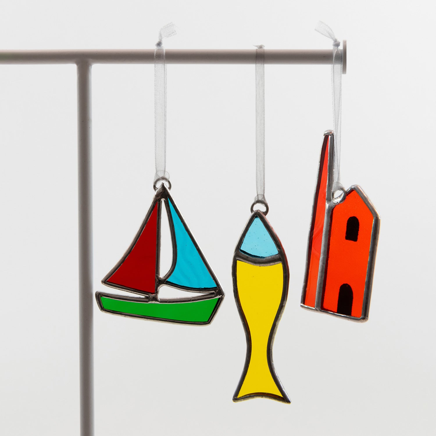 Three glass decorations hanging, on a white background. There's a sailboat, a fish, and an engine house.
