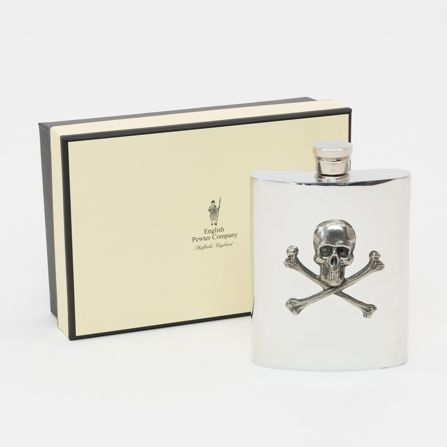 A silver hip flask with a skull and cross bones detail. The hip flask is displayed in front of the presentation gift box.