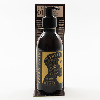 A bottle of beard wash pictured on a white background. It's a dark brown bottle with a white and yellow label. The label features an illustration of a man's face in profile with a large beard.