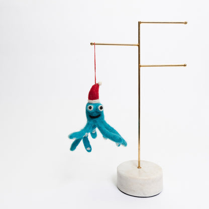 A Christmas decoration pictured on a white background. The decoration is a felt blue octopus wearing a red Christmas hat, smiling.