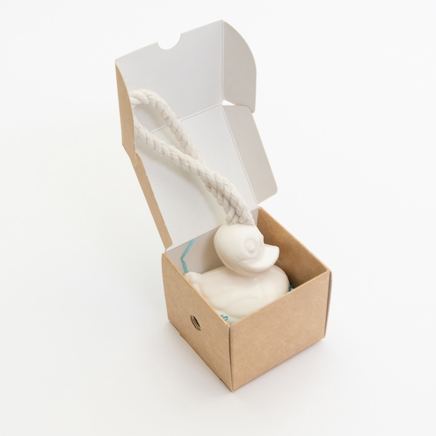 A white duck-shaped soap on a rope in a cardboard gift box. The duck is placed on shredded blue tissue paper in the box. 