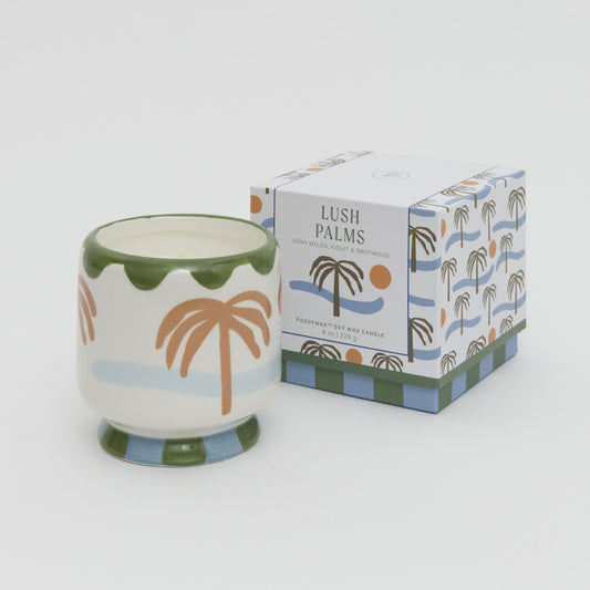 Image of soy candle in a ceramic candle holder painted with palm tree, sea and sun. Candle is placed next to the square box with matching artwork.