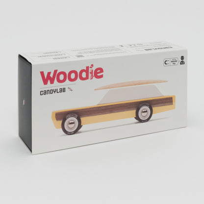 A photograph showing a yellow wooden toy car with wooden surfboard on its roof.