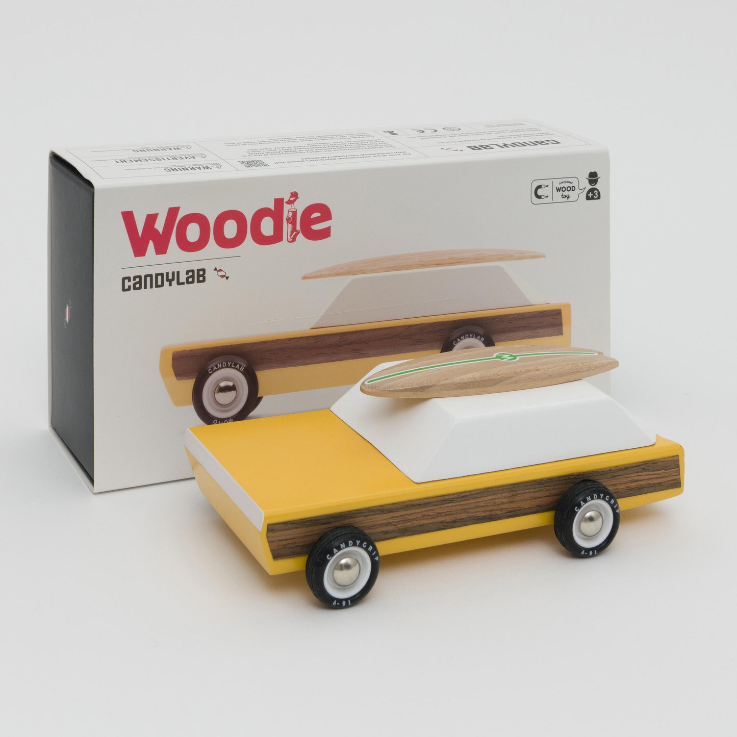A photograph showing a yellow wooden toy car with wooden surfboard on its roof.