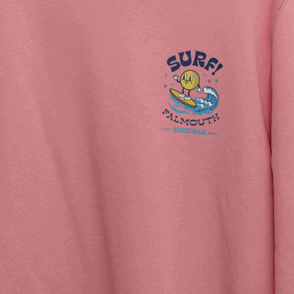 Close up shot of the motif on the front of the bright pink sweatshirt. Motif shows round character surfing a wave with the wording SURF! Falmouth, Cornwall.