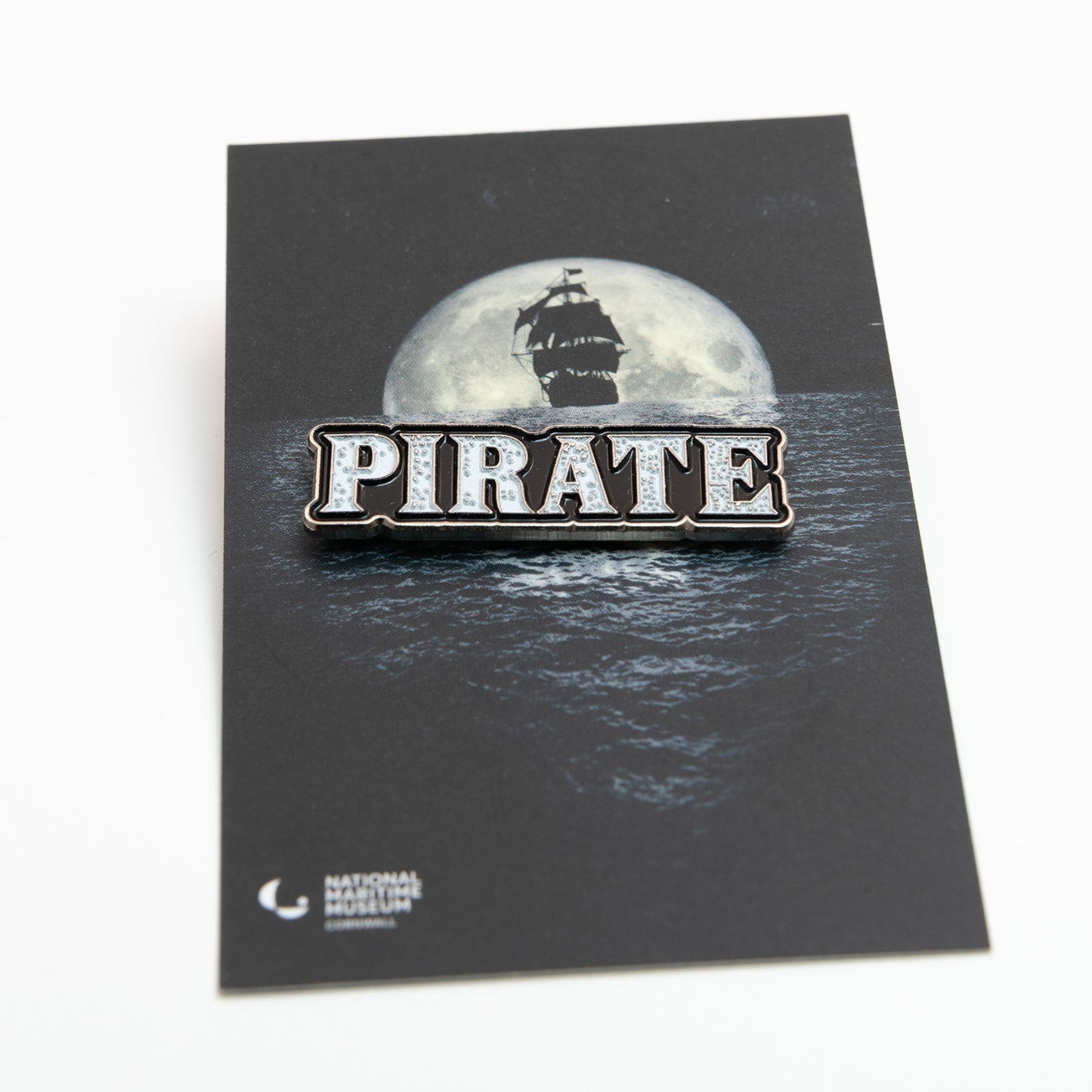 A photo of the National Maritime Museum Cornwall's Pirate Exhibition Pin Badge on branded cardboard, on a white background.