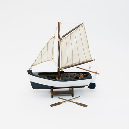 Full view of the model sailing boat with a navy blue and white hull and cream sails. There are two wooden oars on display.