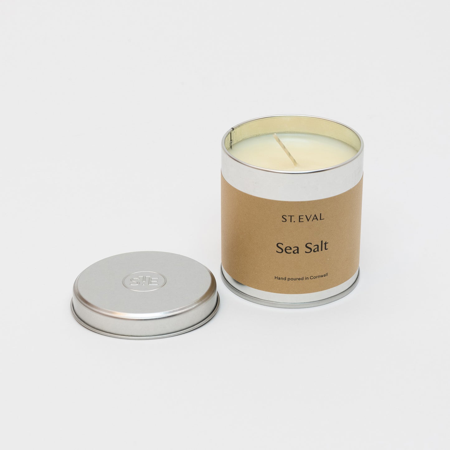 St Eval Sea Salt Candle Tin with tin lid by side.. Plain earthy card label with St. Eval, Sea Salt, Made in Cornwall. Cream white candle with standing wick.