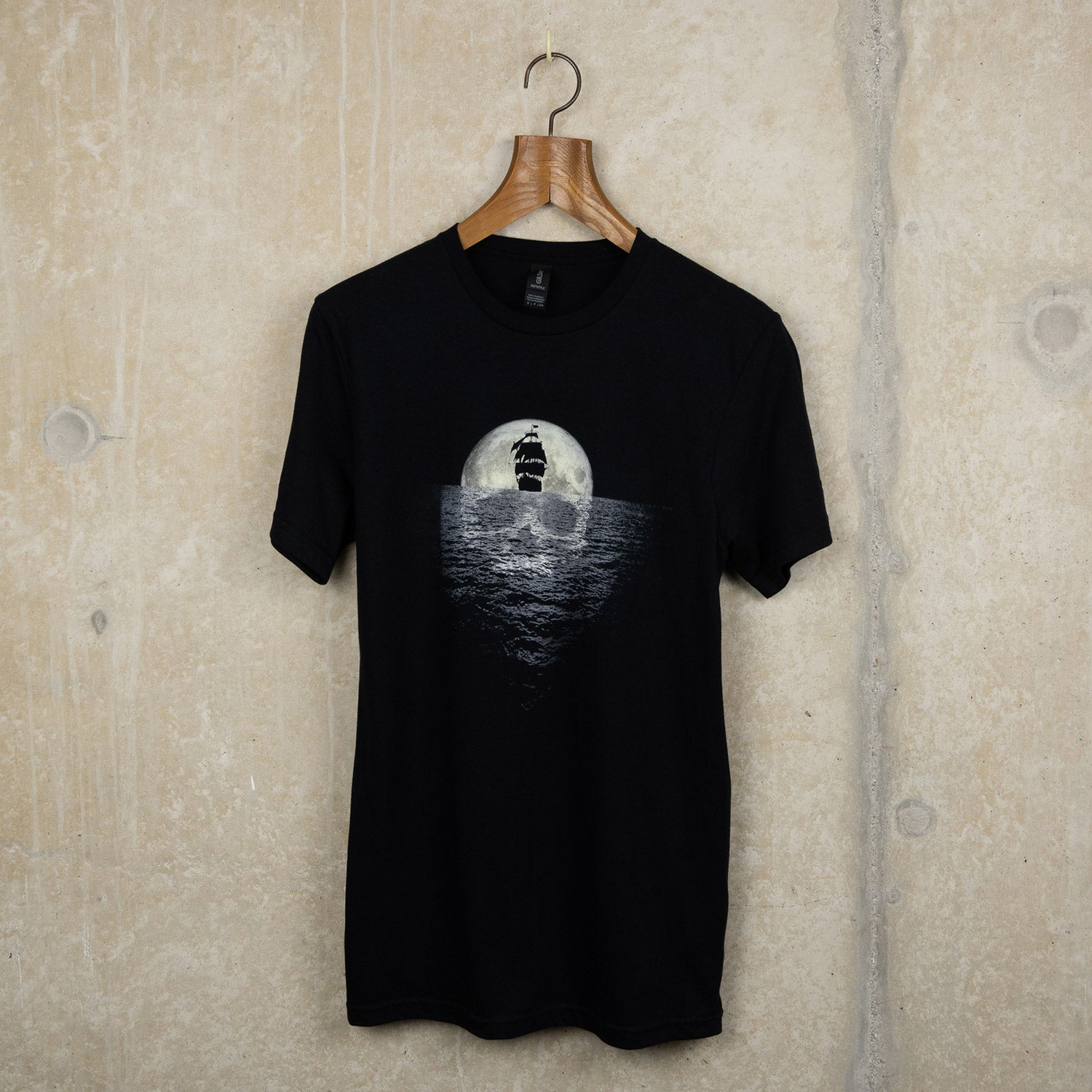 Black t-shirt with pirate exhibition artwork of a sailing ship sailing away from a full moon reflecting a skull in the sea
