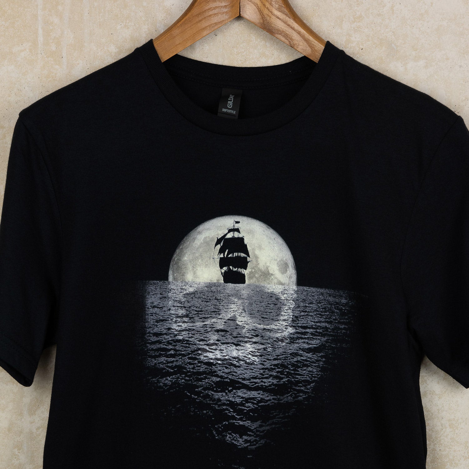 Black t-shirt with pirate exhibition artwork of a sailing ship sailing away from a full moon reflecting a skull in the sea, close up