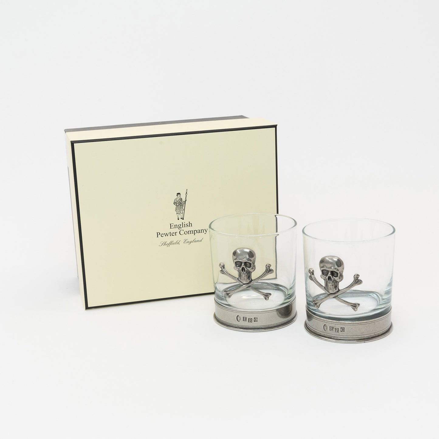 Two glass tumblers with a pewter base and a pewter skull and crossbones on the side of the glass. The glasses are in front of a cream coloured gift presentation box.