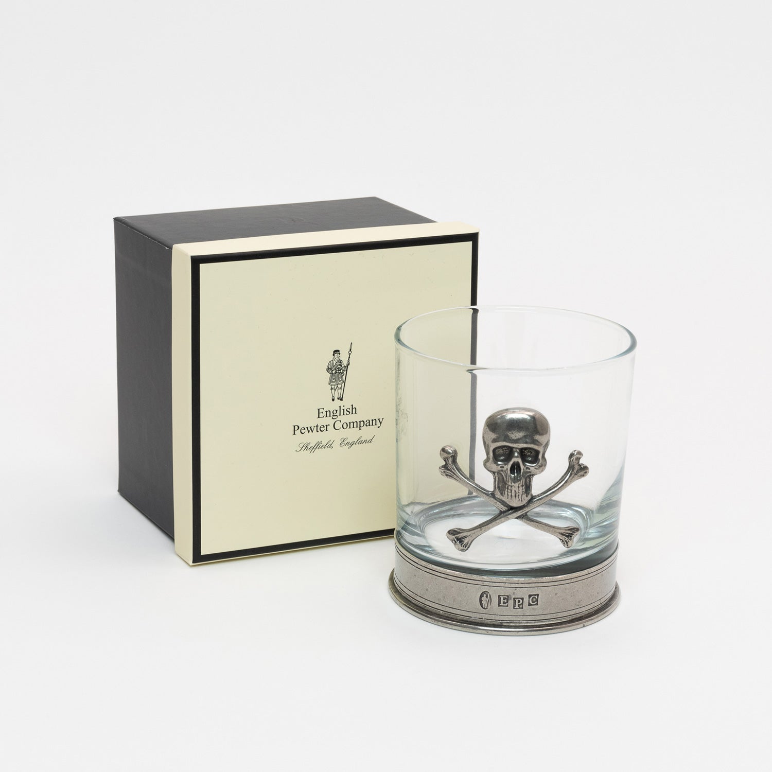 Clear glass with a pewter base and pewter skull and cross bones on the side of the tumbler. The glass is next to the cream coloured gift box.