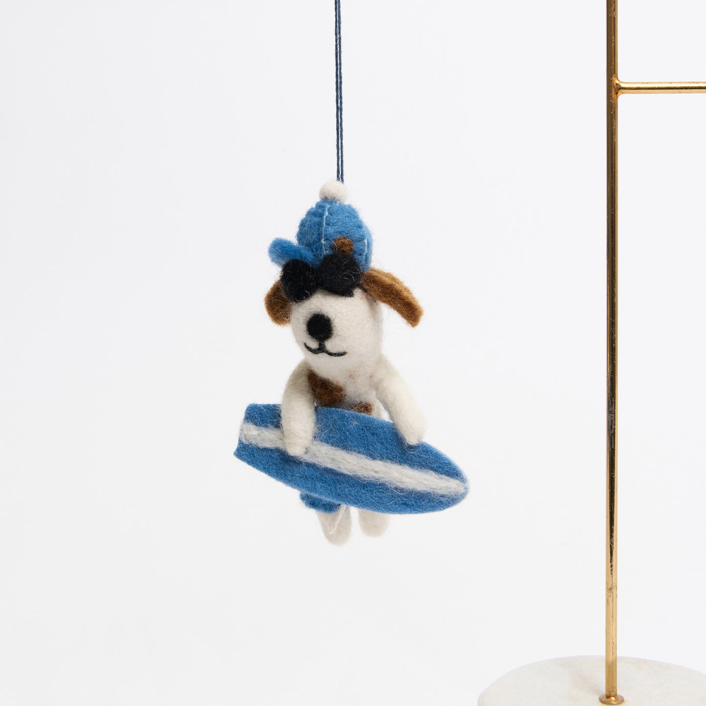 A surfer dog felt decoration pictured on a white background. The dog is white and brown, is wearing a blue hat and blue shorts, black sunglasses, and holding a blue and white surfboard. 