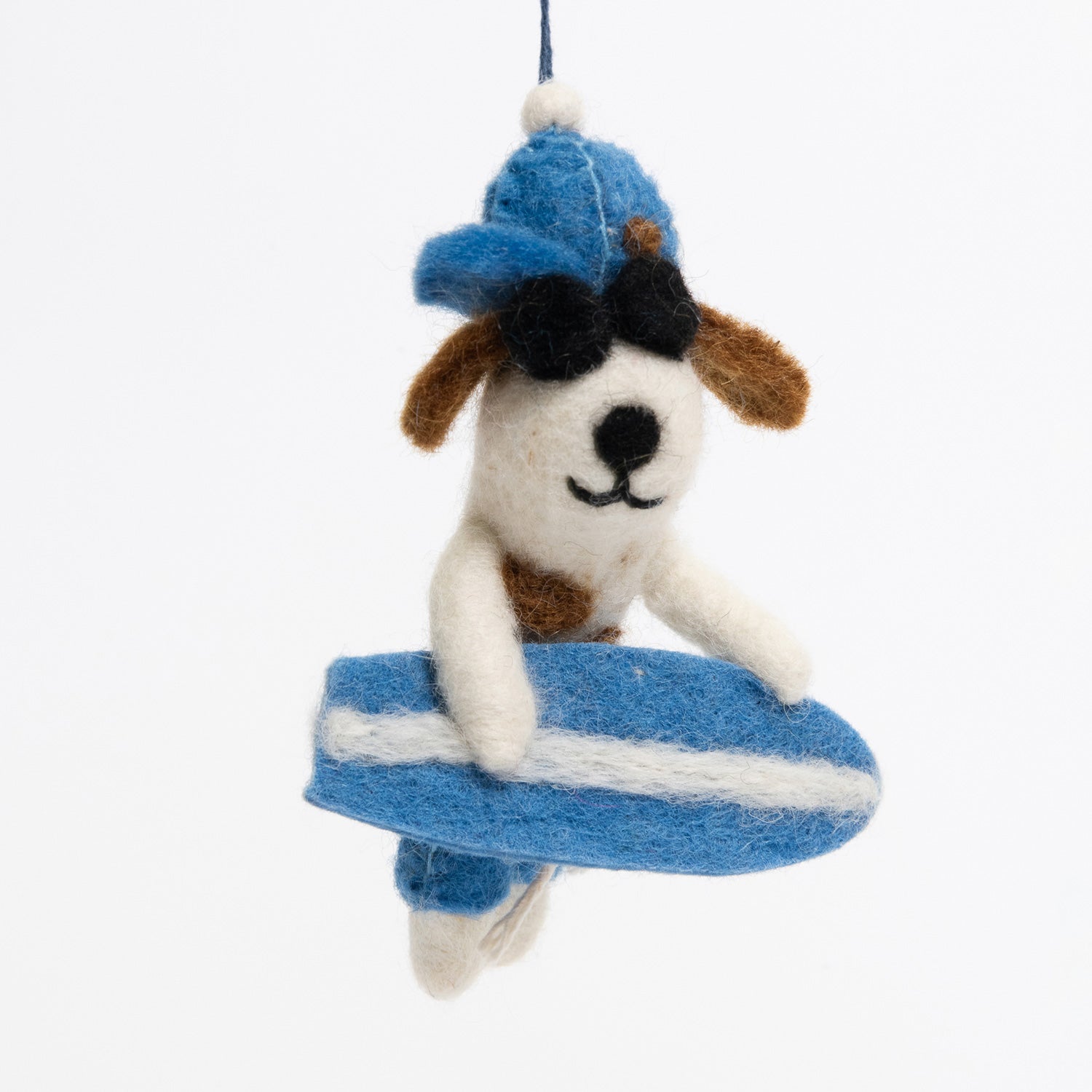 A surfer dog felt decoration pictured on a white background. The dog is white and brown, is wearing a blue hat and blue shorts, black sunglasses, and holding a blue and white surfboard. 