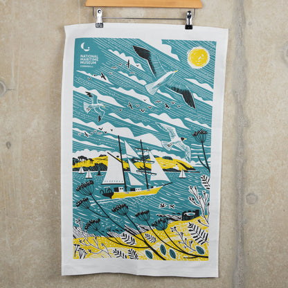 Tall ships teal towel with an illustration of a tall ship in blue, white and yellow. The tea towel is hanging from a clip hanger against a light grey background.