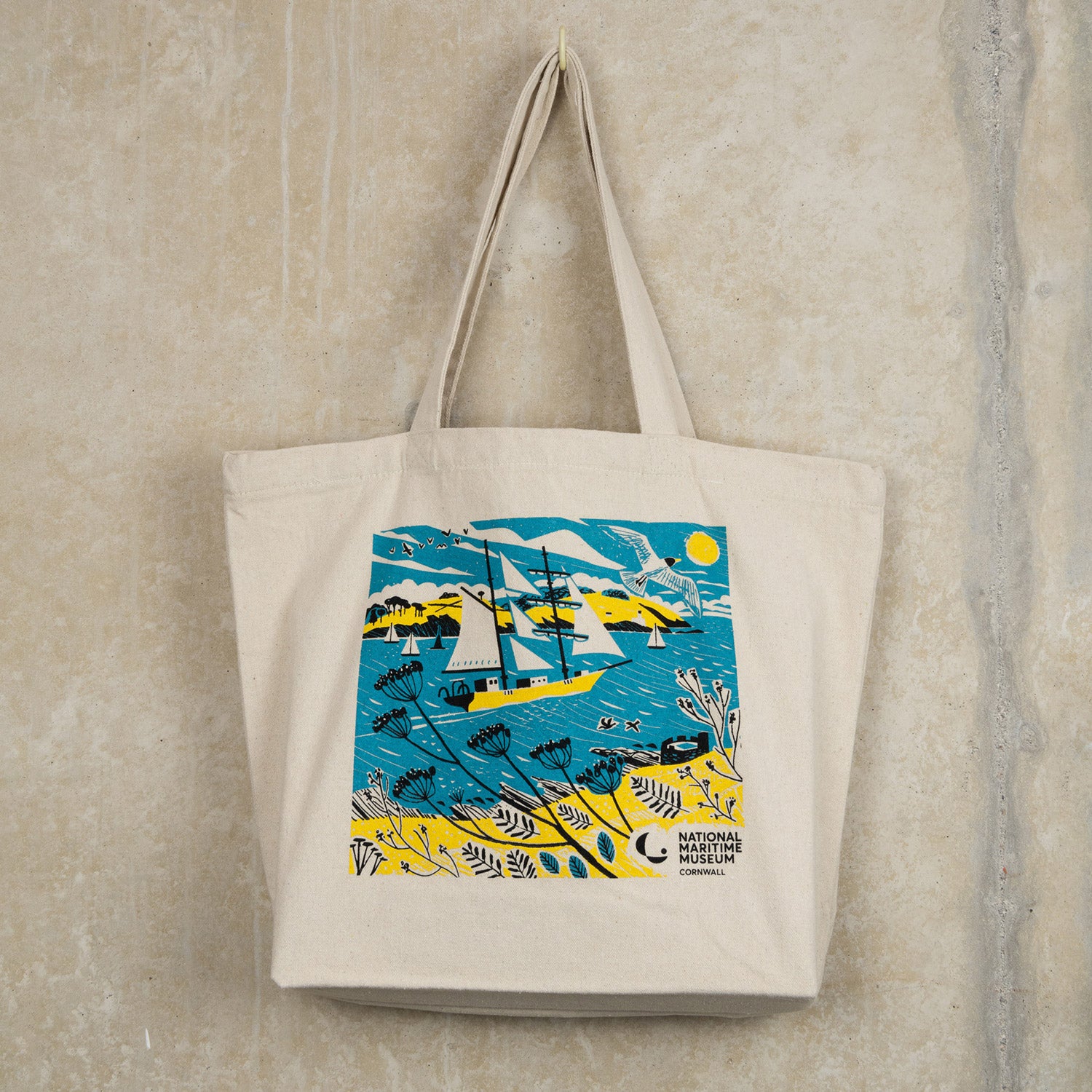 The Falmouth Tall Ship Tote Bag featuring a blue, yellow and white design of a tall ship in full sail. The tote bag is hanging from a hook against a light grey background.