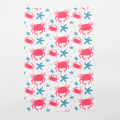 The crab and starfish tea towel features pink crabs and blue starfish on a light cream background. The tea towel is against a white background.