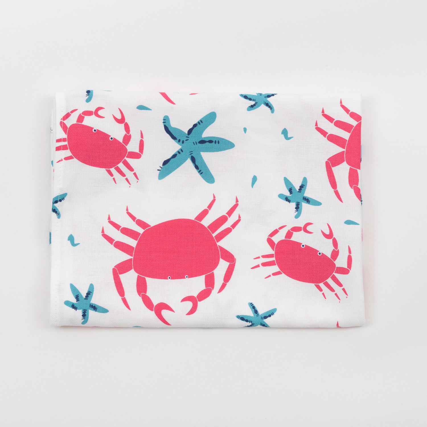 The crab and starfish tea towel features pink crabs and blue starfish on a light cream background and is folded against a white background.
