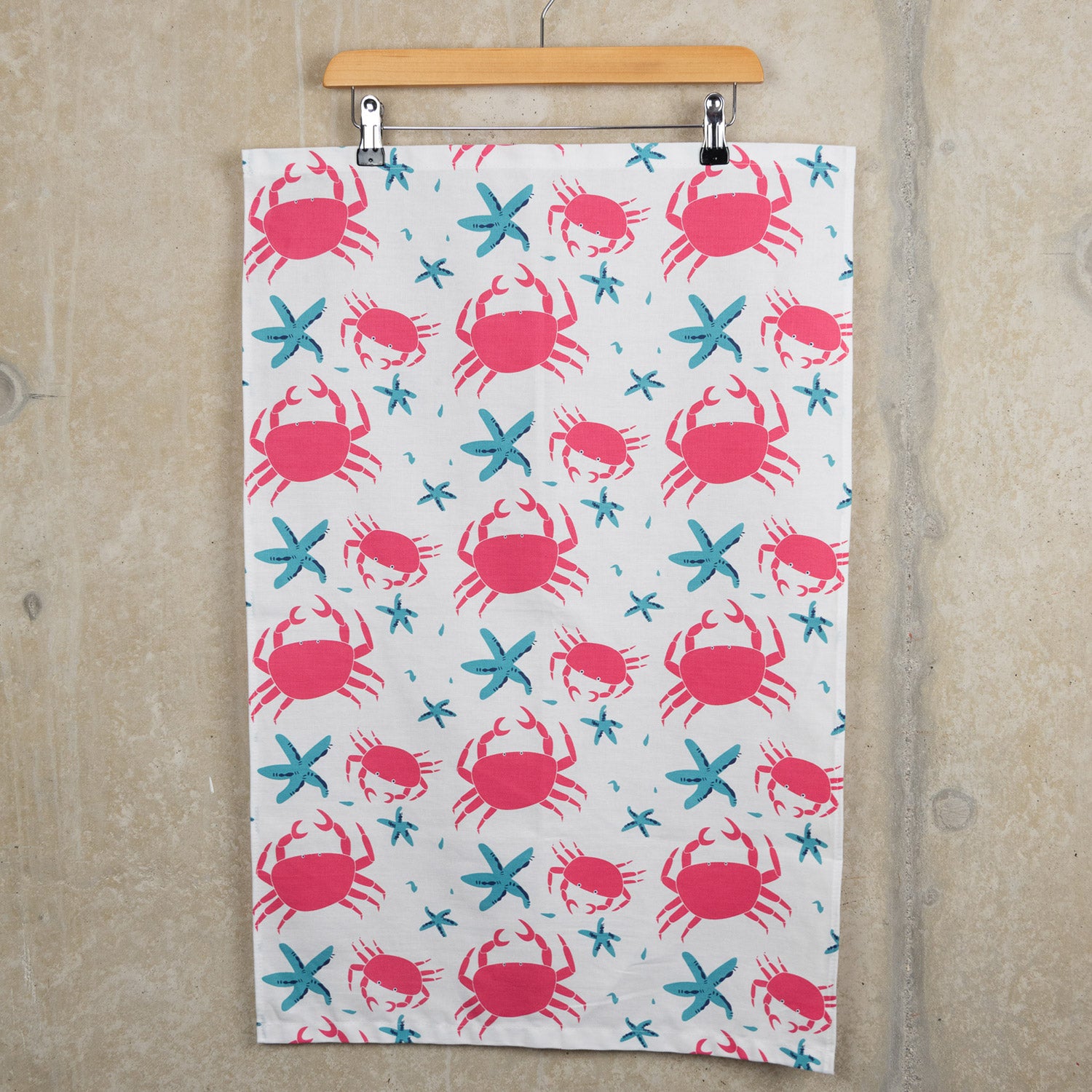 The crab and starfish tea towel features pink crabs and blue starfish on a light cream background. The tea towel is hanging from a clip hanger against a light grey background.