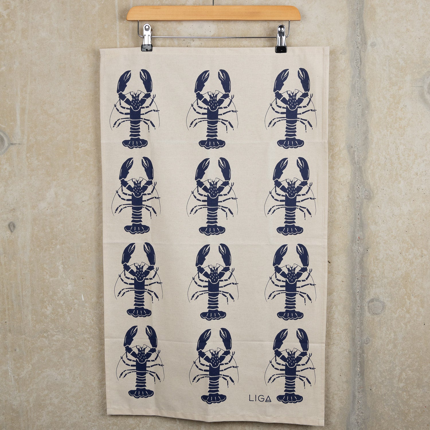Navy lobster repeated pattern on a cream coloured tea towel. The Navy Lobster Tea Towel is hanging from a clip hanger against a grey background.