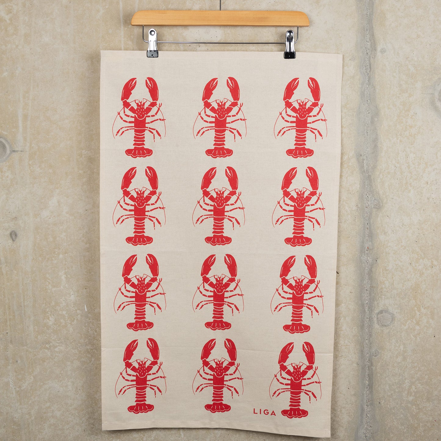Red lobster repeating pattern on a cream coloured back ground. The Red Lobster Tea Towel is hanging from a clip hanger on a grey background.