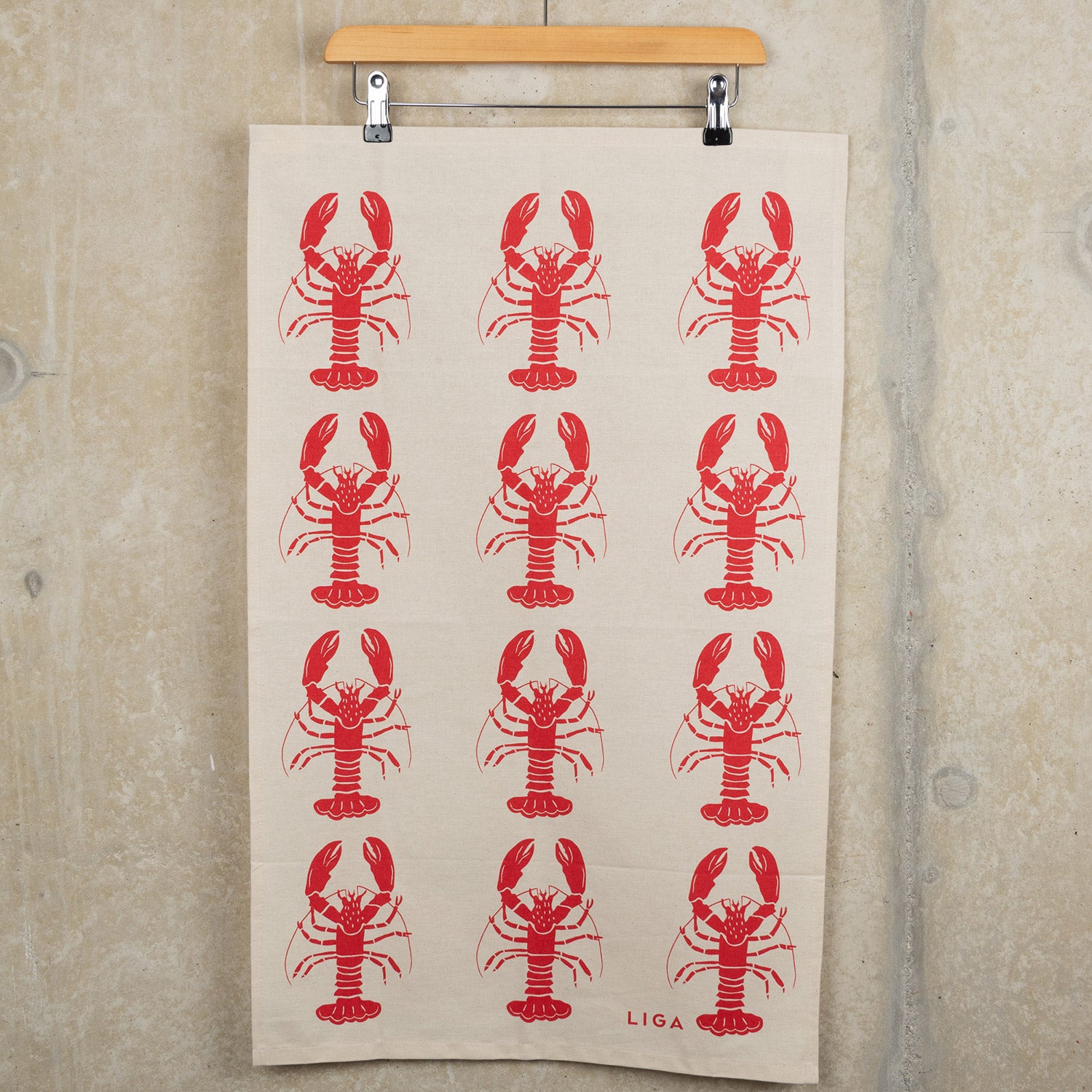 Red lobster repeating pattern on a cream coloured back ground. The Red Lobster Tea Towel is hanging from a clip hanger on a grey background.