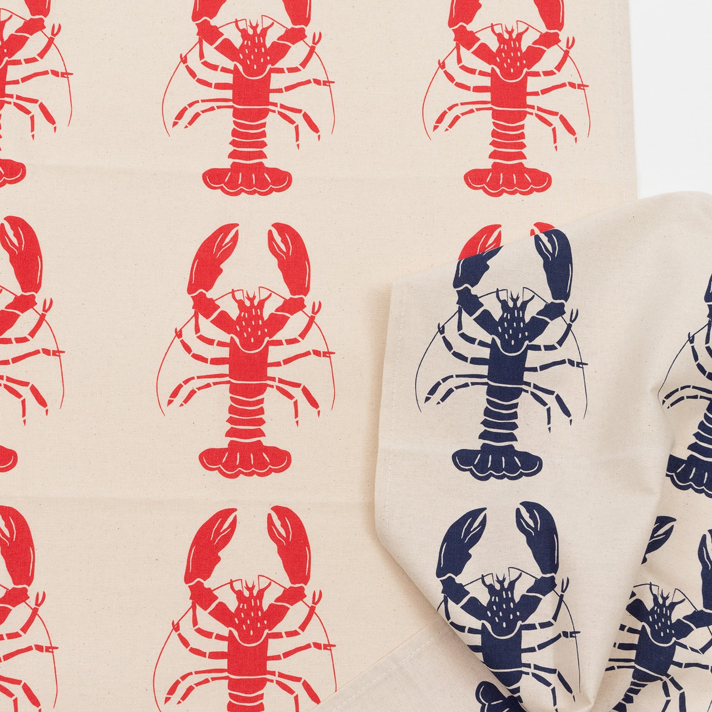 Red lobster repeating pattern on a cream coloured back ground. The Red Lobster Tea Towel is overlaid by the navy version of the tea towel.