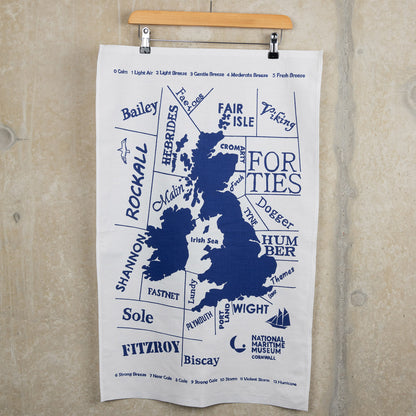 Blue and white shipping forecast tea towel hanging from a hanger on a grey background.